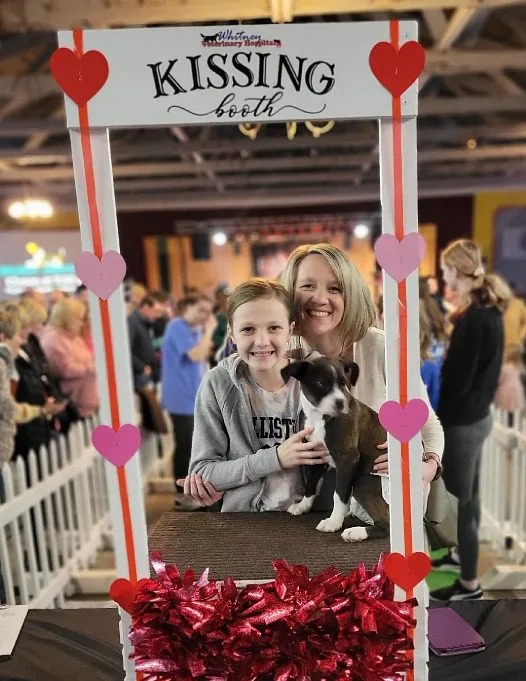Picture of a woman and young girl posing with a puppy in front of a kissing booth